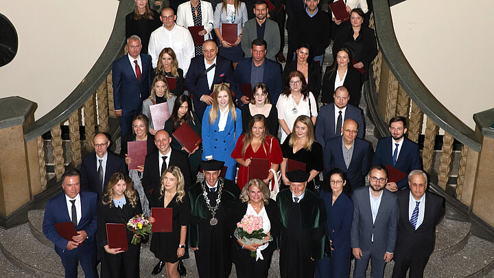 Odświętnie ubrani absolwenci studiów podyplomowych stojący na schodach w hallu głównym budynku AO pozujący z dyplomami