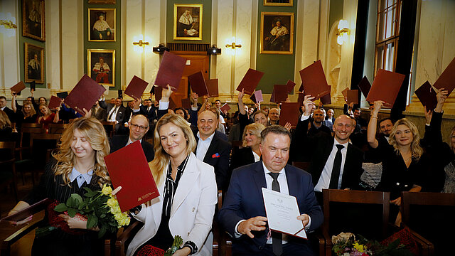 Odświętnie ubrani ludzie trzymający w rękach okładki dyplomów w auli budynku A0.