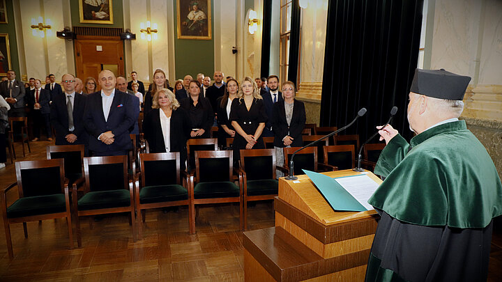 Widoczne plecy kierownika studiów w birecie i todze, stojącego przodem do mównicy, a na drugim planie widoczni absolwenci studiów w auli głównej budynku A0